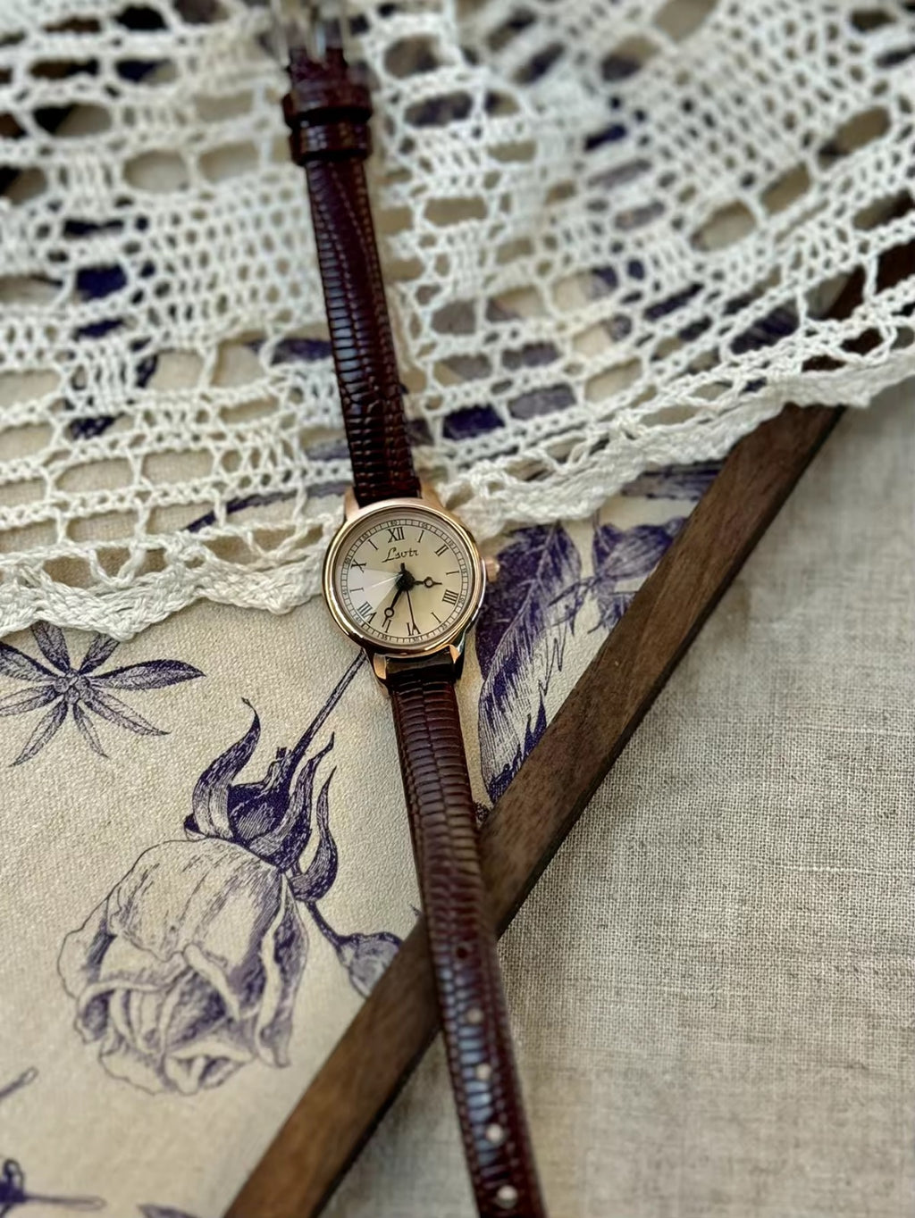 Women’s rose gold Roman numeral watch with brown lizard-grain
leather strap on linen background