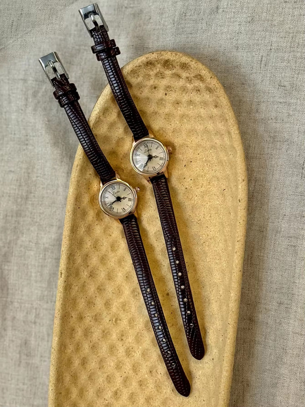 Women’s rose gold Roman numeral watch with brown lizard-grain
leather strap on linen background