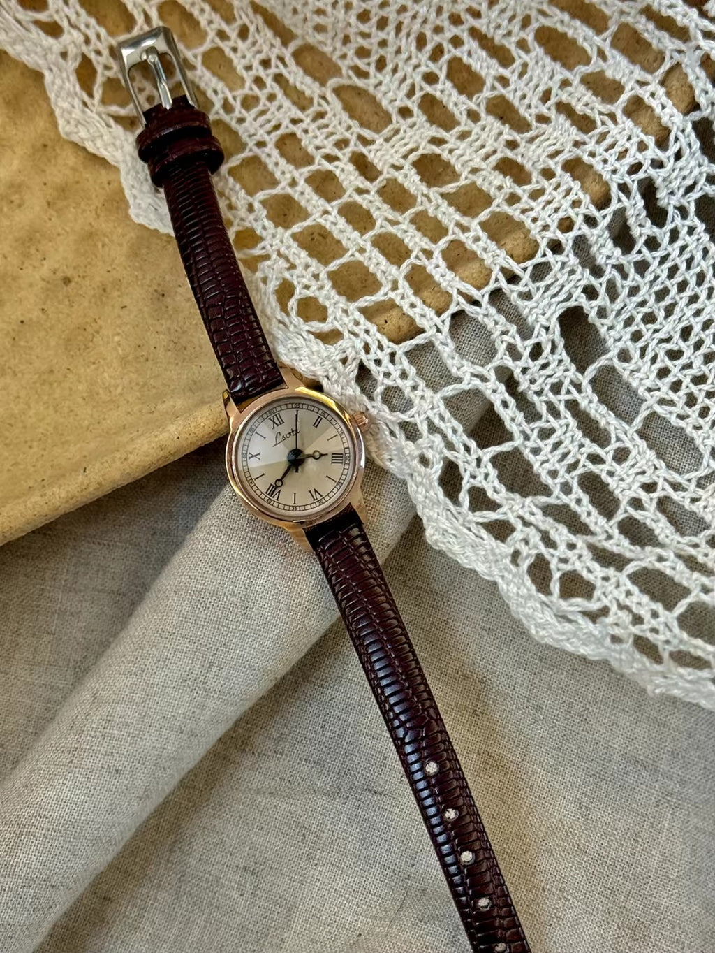 Women’s rose gold Roman numeral watch with brown lizard-grain
leather strap on linen background
