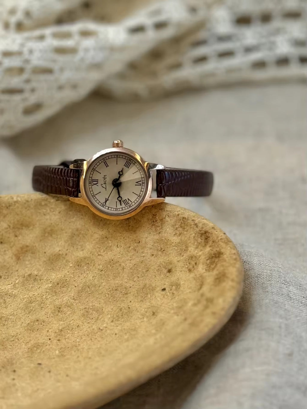 Women’s rose gold Roman numeral watch with brown lizard-grain
leather strap on linen background