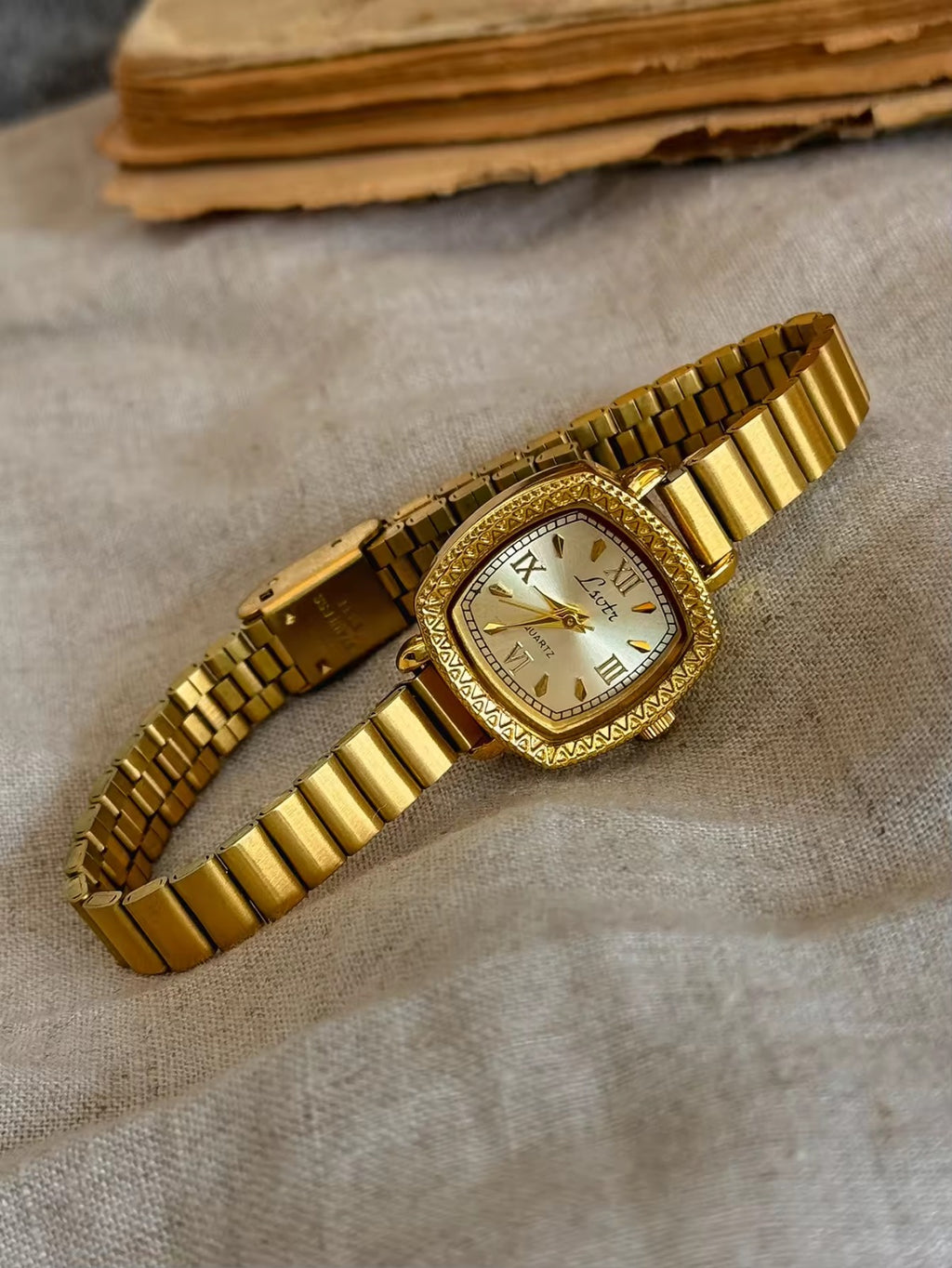 Gold cushion square watch with deeply engraved geometric chevron
bezel, Roman numeral and baton marker champagne dial and structured gold expansion flex bracelet for women by Le Belle Heure