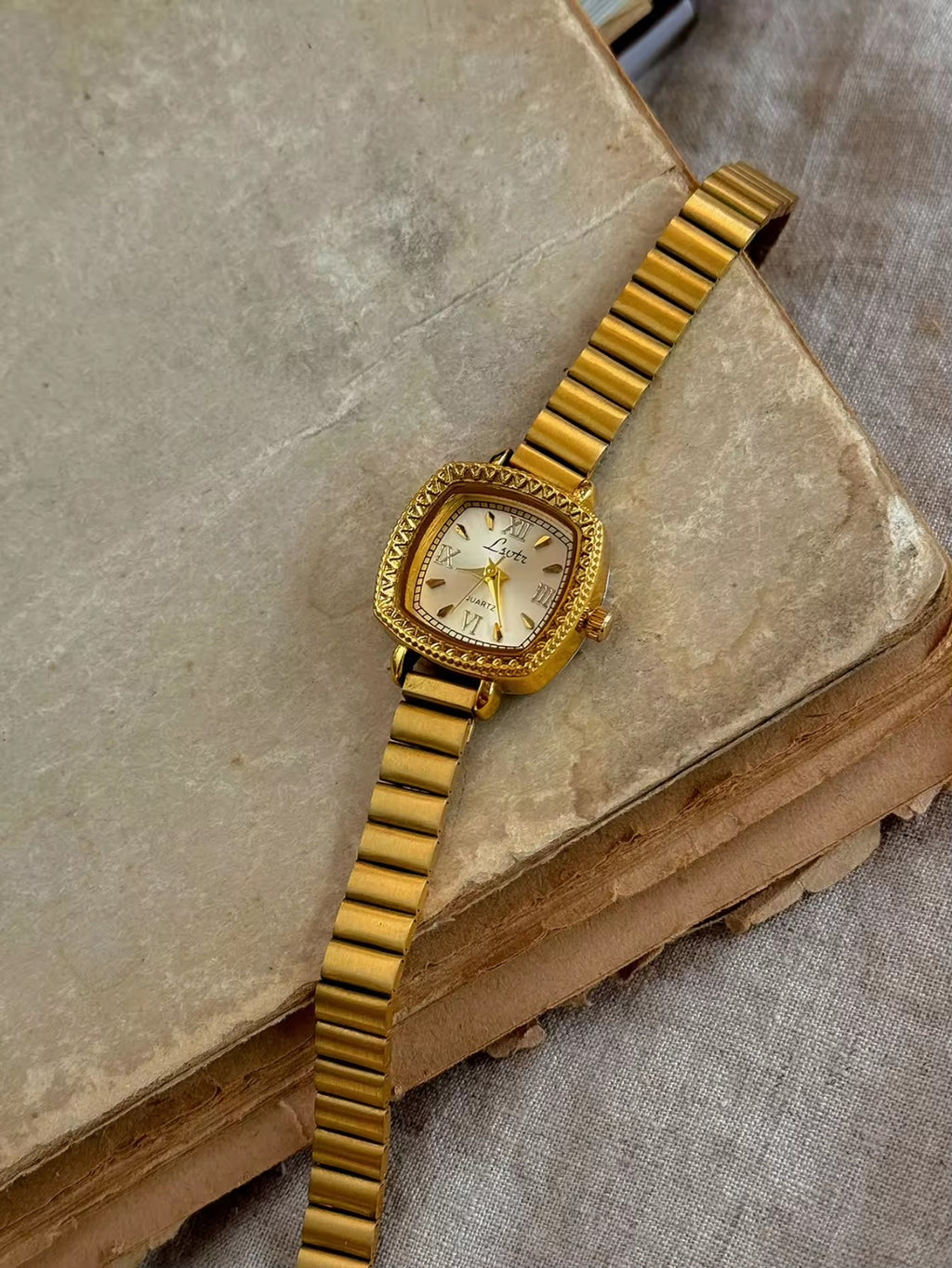 Gold cushion square watch with deeply engraved geometric chevron
bezel, Roman numeral and baton marker champagne dial and structured gold expansion flex bracelet for women by Le Belle Heure