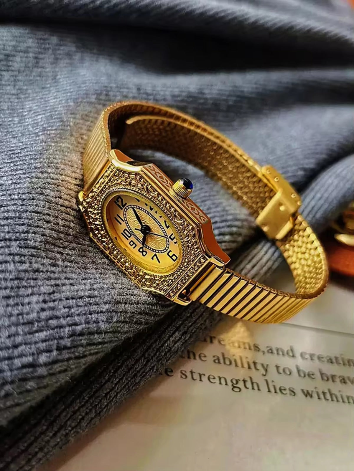 Two women’s vintage gold octagonal watches with engraved filigree bezels, white Arabic numeral dials, and gold accordion ribbed flex bracelets on an antique book — Le Belle Heure
