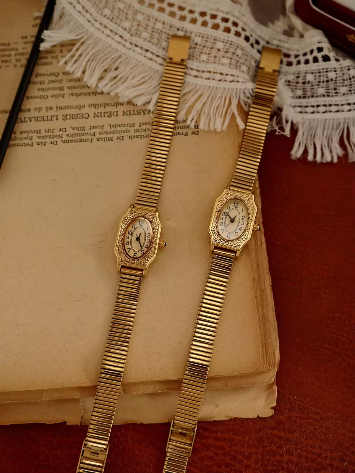 Two women’s vintage gold octagonal watches with engraved filigree bezels, white Arabic numeral dials, and gold accordion ribbed flex bracelets on an antique book — Le Belle Heure