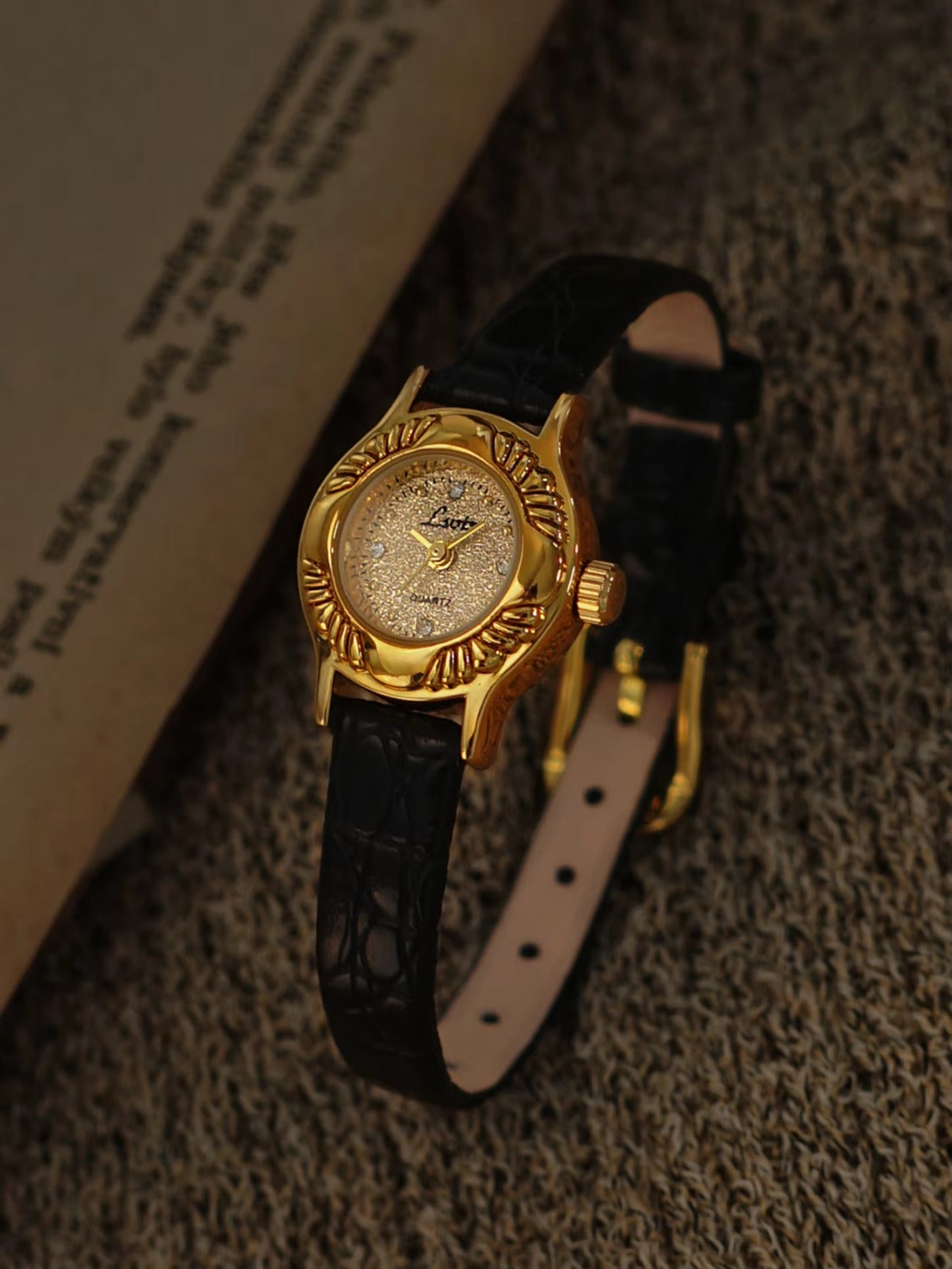 Gold oval watch with deeply engraved feather and leaf floral case,
champagne glitter dial, sculpted gold lugs and slim black crocodile texture leather strap for women by Le Belle Heure