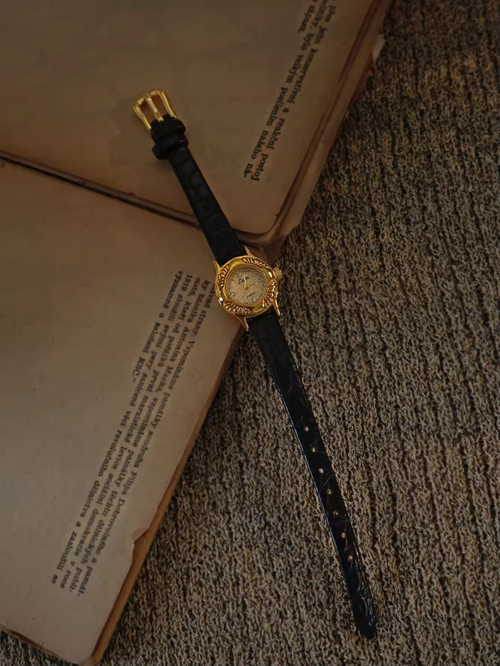 Gold oval watch with deeply engraved feather and leaf floral case,
champagne glitter dial, sculpted gold lugs and slim black crocodile texture leather strap for women by Le Belle Heure