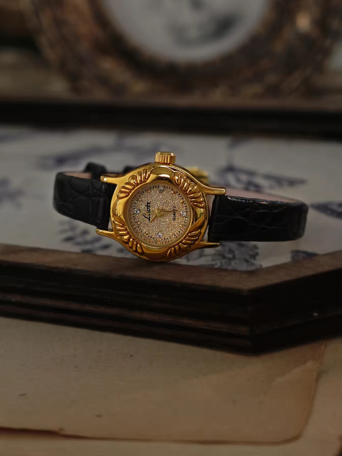 Gold oval watch with deeply engraved feather and leaf floral case,
champagne glitter dial, sculpted gold lugs and slim black crocodile texture leather strap for women by Le Belle Heure