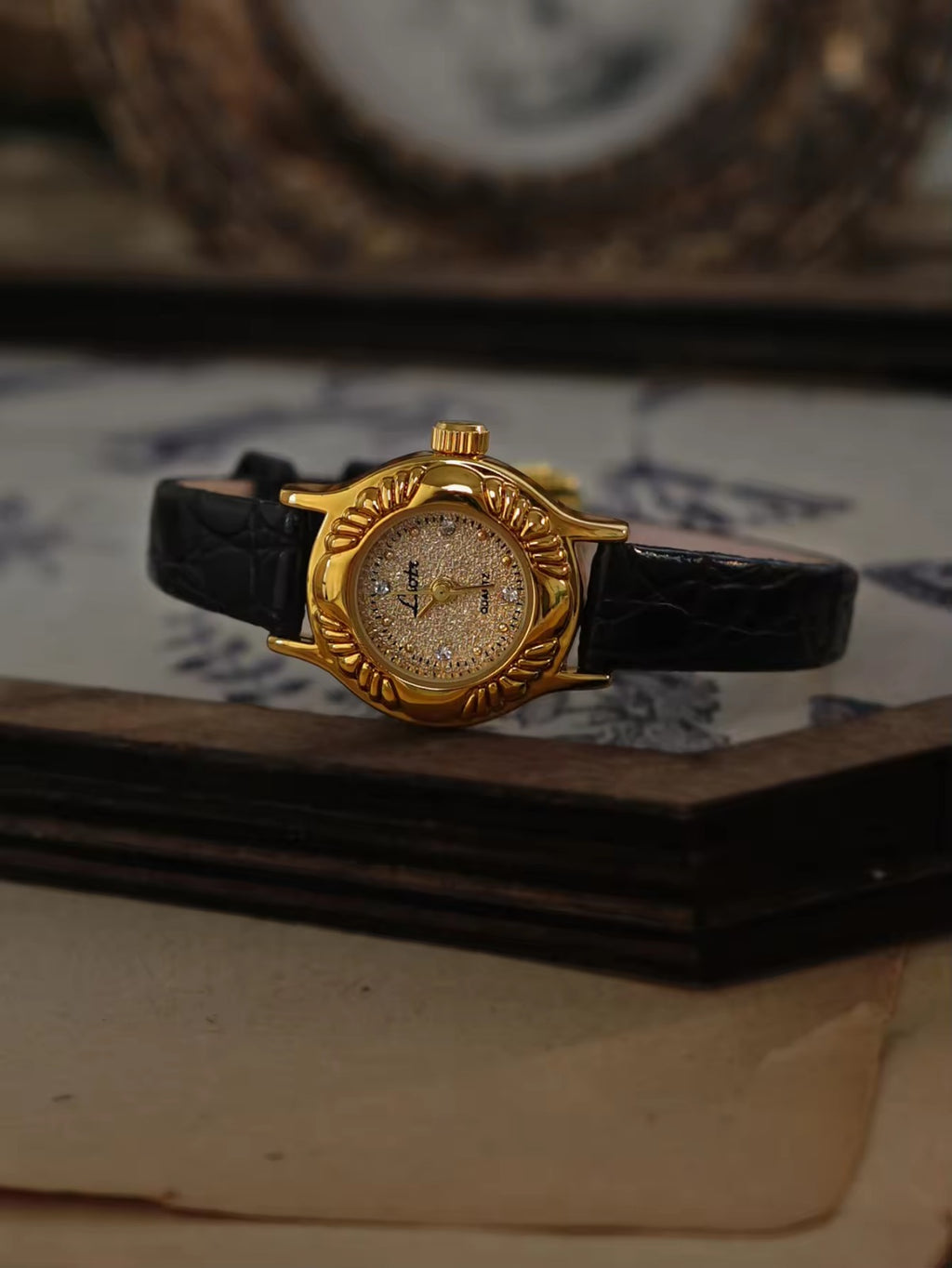 Gold oval watch with deeply engraved feather and leaf floral case,
champagne glitter dial, sculpted gold lugs and slim black crocodile texture leather strap for women by Le Belle Heure
