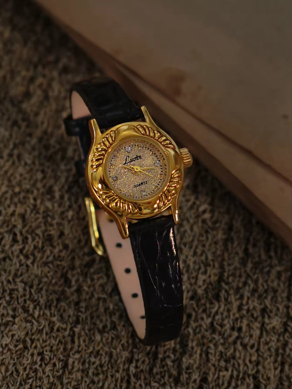Gold oval watch with deeply engraved feather and leaf floral case,
champagne glitter dial, sculpted gold lugs and slim black crocodile texture leather strap for women by Le Belle Heure